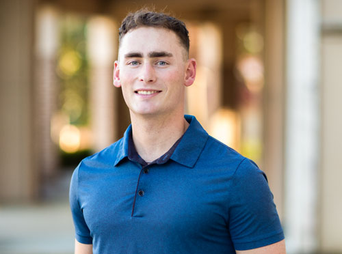 Professional portrait of a young man in a blue polo shirt standing outdoors, representing business leadership and AI consulting services.