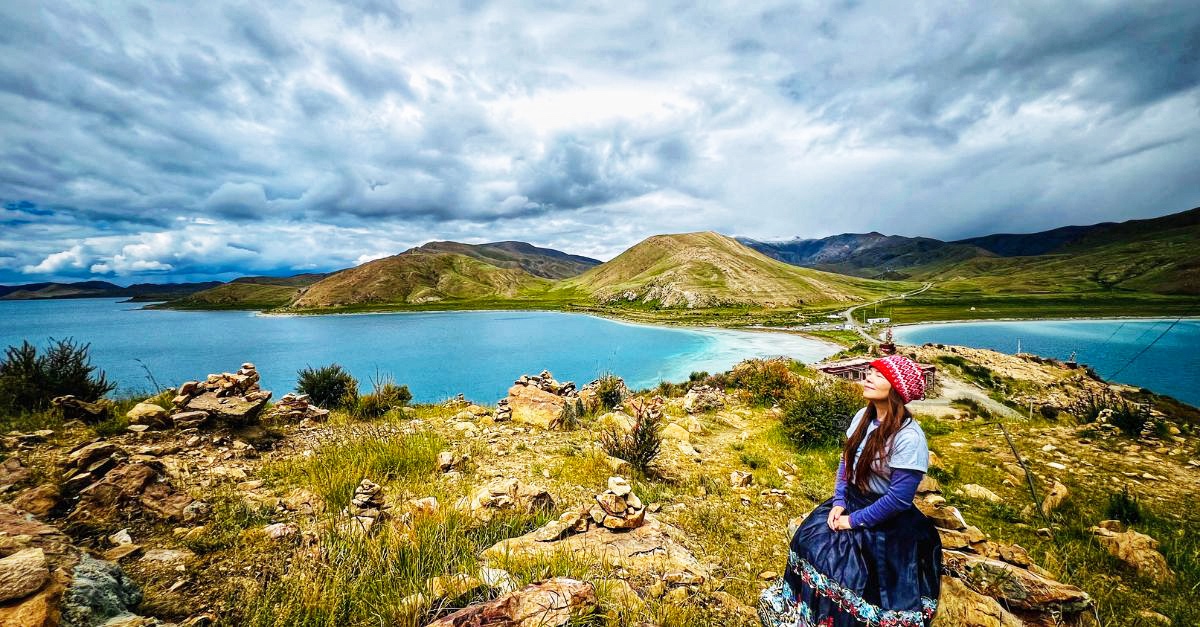Riya Wang seated beside a blue lake in Tibet beneath a dramatic sky