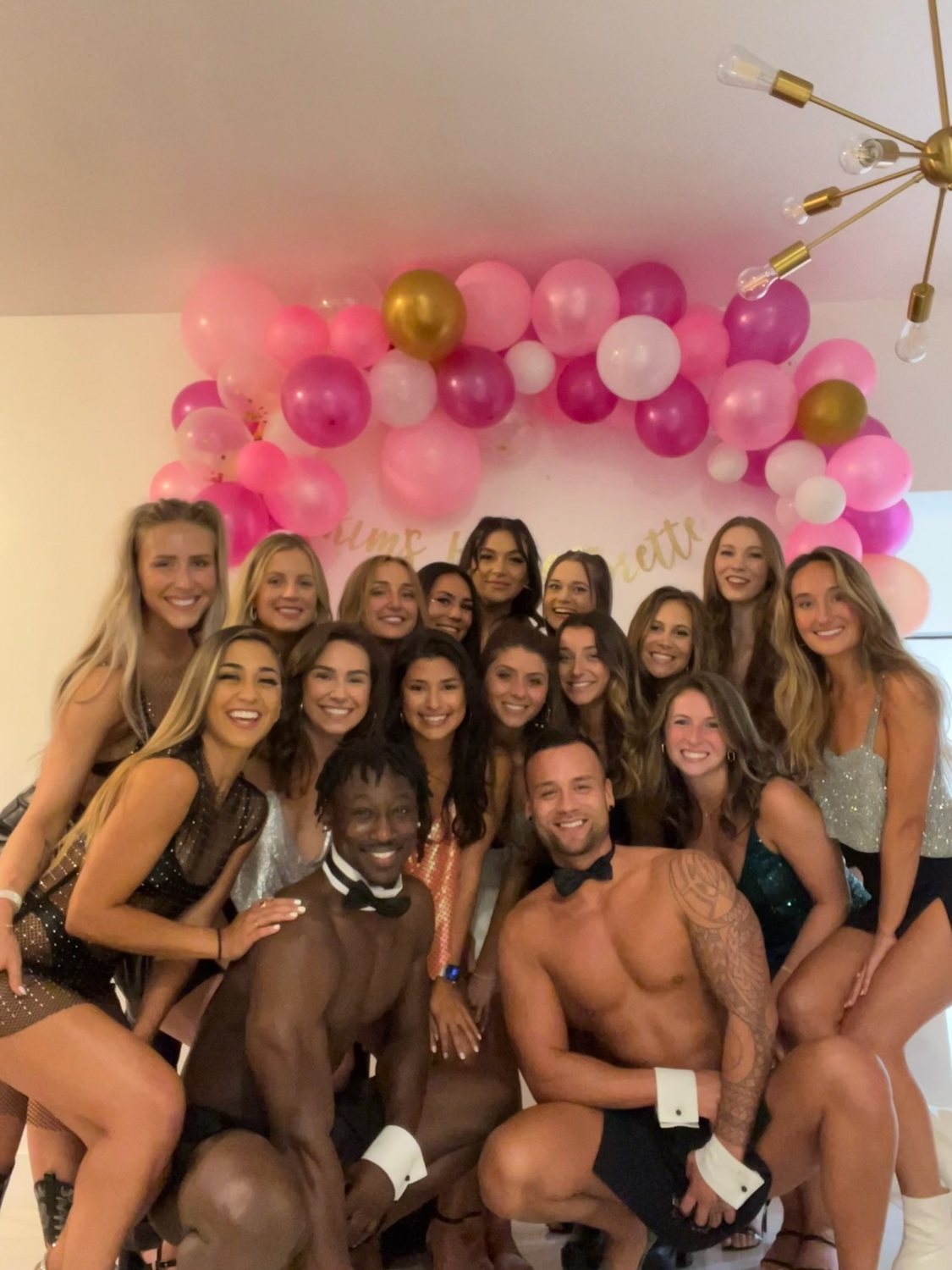 A bachelorette group celebrating under a pink balloon arch with two Cheeky Butlers