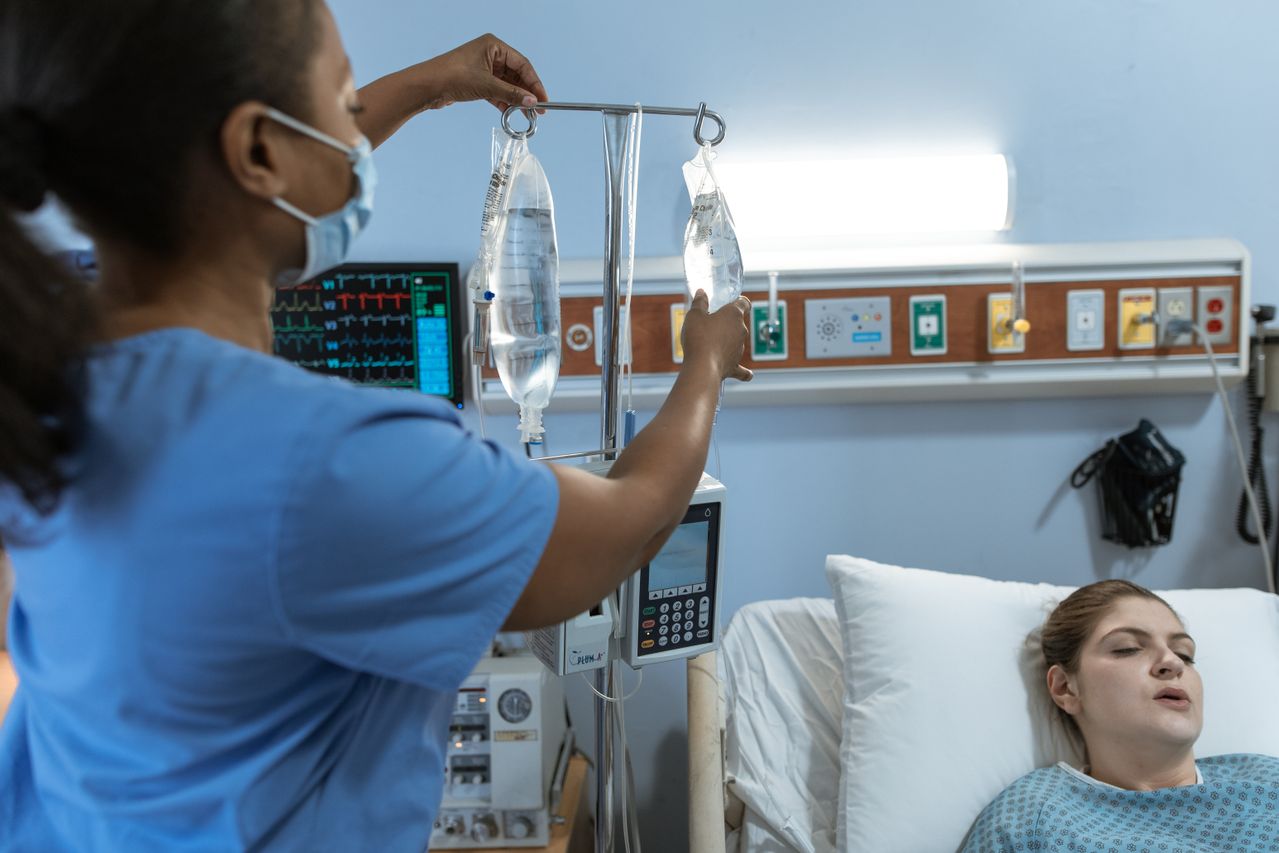 A nurse in a hospital setting adjusts an IV drip for a patient lying in bed, showcasing medical care. A nurse in a hospital setting adjusts an IV drip for a patient lying in bed, showcasing medical care.