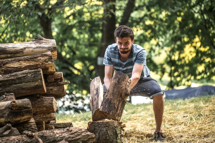 A man in a patterned t-shirt splitting a log with an axe next to a large pile of stacked firewood, highlighting professional wood preparation services by SGJ Landscaping in Honesdale, PA.