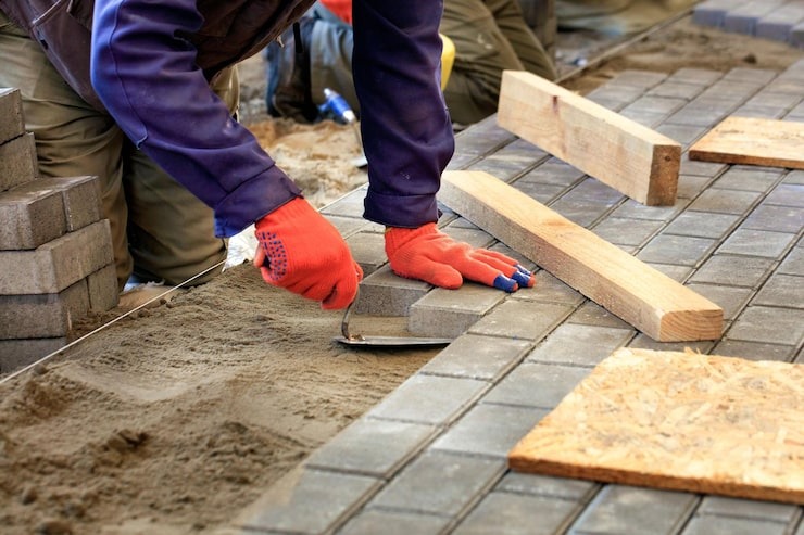 A professional paver installer wearing orange safety gloves, using a trowel to expertly lay bricks on a sand base for a custom patio, by SGJ Landscaping in Honesdale, PA.