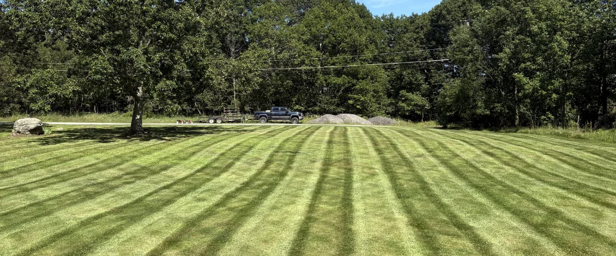 A freshly mowed lawn with striped patterns in the foreground, and a truck with a trailer parked in the background.