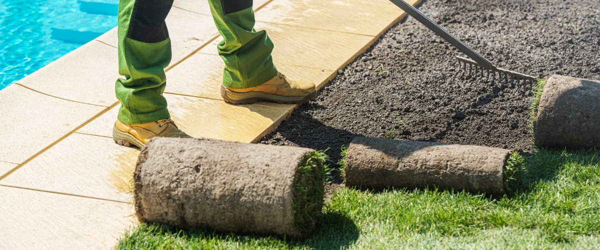 A gardener in green trousers and work boots lays rolls of sod beside a swimming pool on a sunny day. A gardener in green trousers and work boots lays rolls of sod beside a swimming pool on a sunny day.
