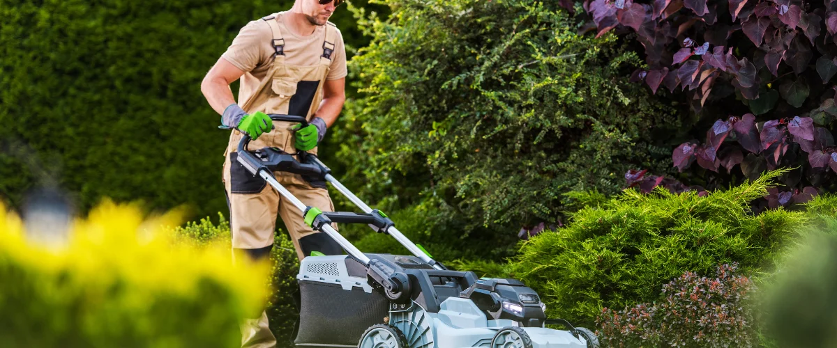 A man in tan overalls and green gloves operates a modern lawnmower in a lush green and purple garden. A man in tan overalls and green gloves operates a modern lawnmower in a lush green and purple garden.