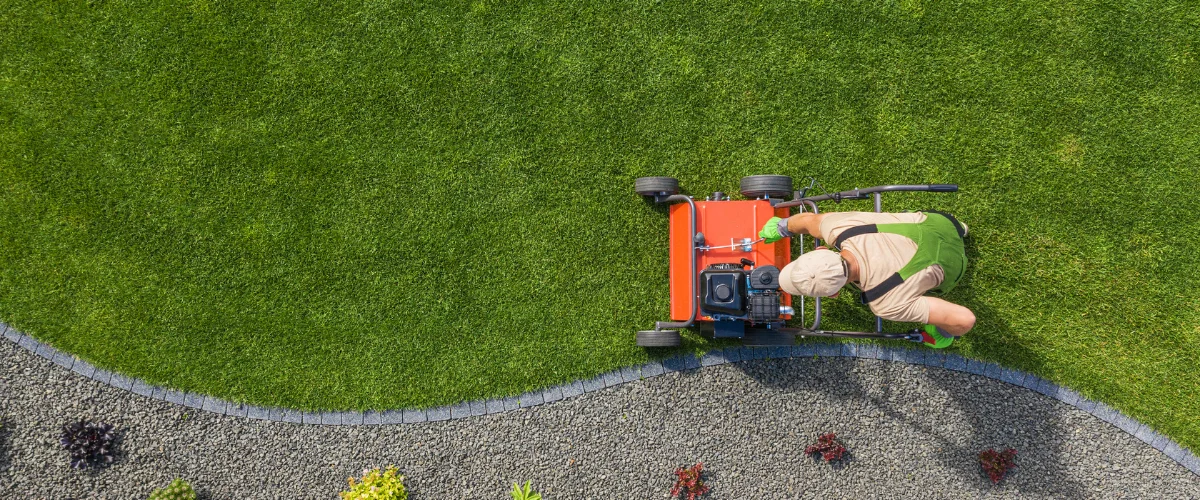 An overhead shot of a man in tan and green operating an orange lawn aerator on a vibrant green lawn next to a gravel path. An overhead shot of a man in tan and green operating an orange lawn aerator on a vibrant green lawn next to a gravel path.