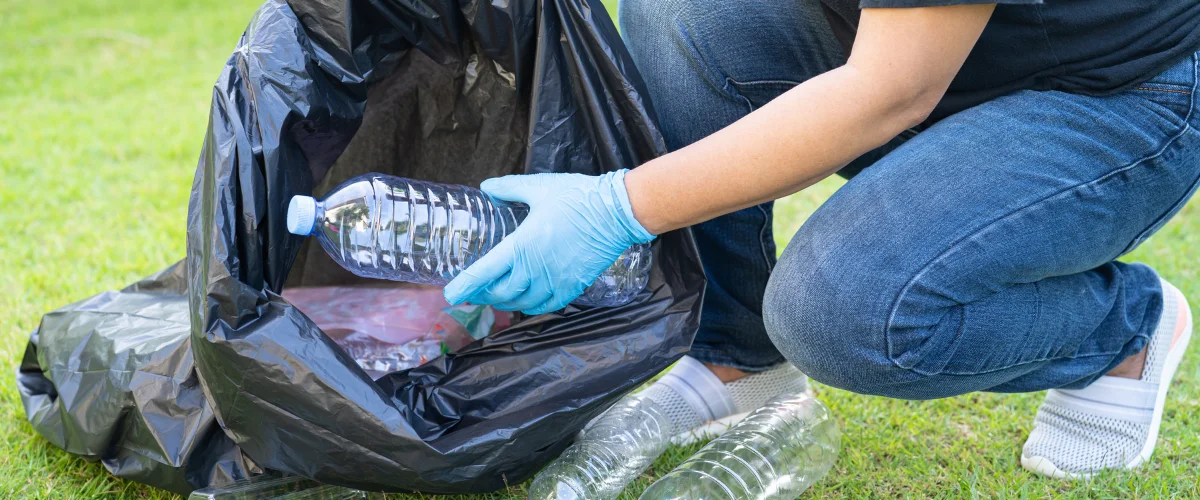 A person in blue gloves and jeans crouches on grass, putting a plastic bottle into a large black trash bag. A person in blue gloves and jeans crouches on grass, putting a plastic bottle into a large black trash bag.