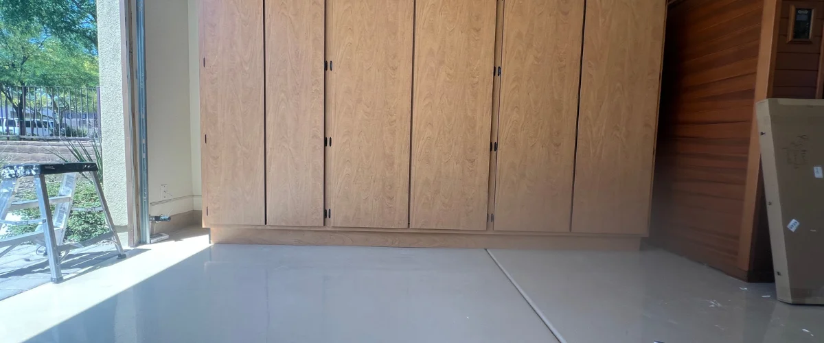 A light wood cabinet system against a wall inside a garage with gray epoxy flooring and an open door showing outside.