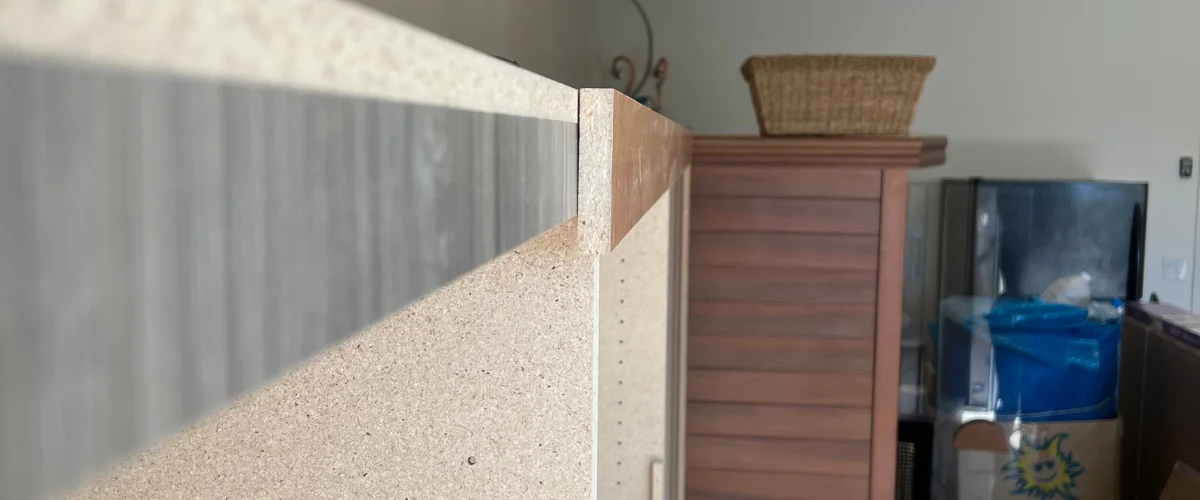 Close-up view of a particulate board shelf with an unfinished edge, positioned above a wooden cabinet and wicker basket.