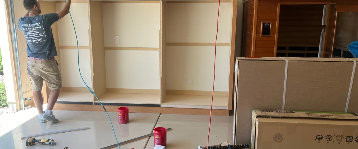 A man in a blue t-shirt and tan shorts installing light wood cabinets in a garage with tools on the floor.