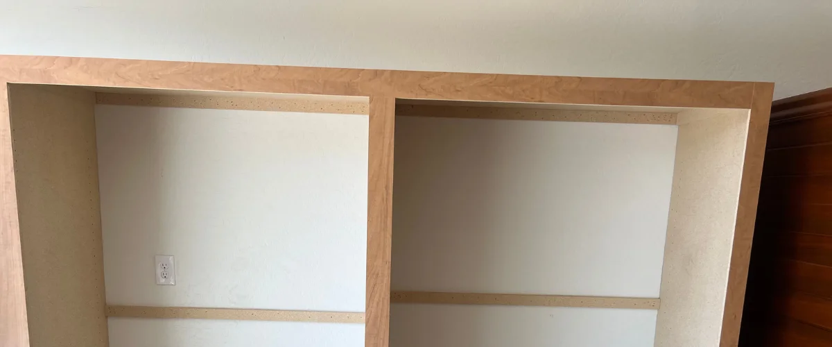 A close-up of light-colored wooden shelves being installed, with a white electrical outlet visible on the back wall.