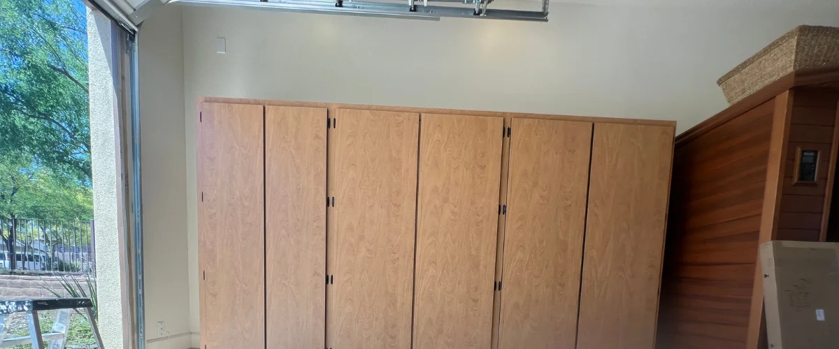 Light wood cabinets filling much of a garage wall, near a side door open to an outside view of trees and sunlight.