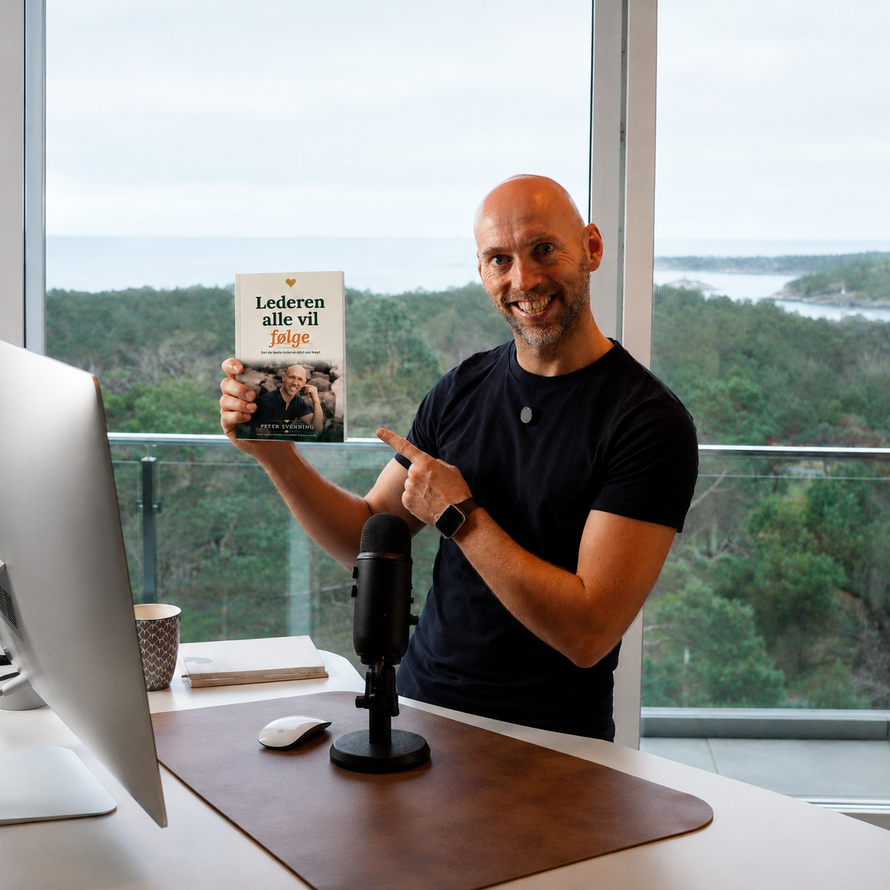Peter Svenning ved skrivebordet sitt, holder fram boka «Lederen alle vil følge» foran et stort vindu med utsikt over skog og hav.