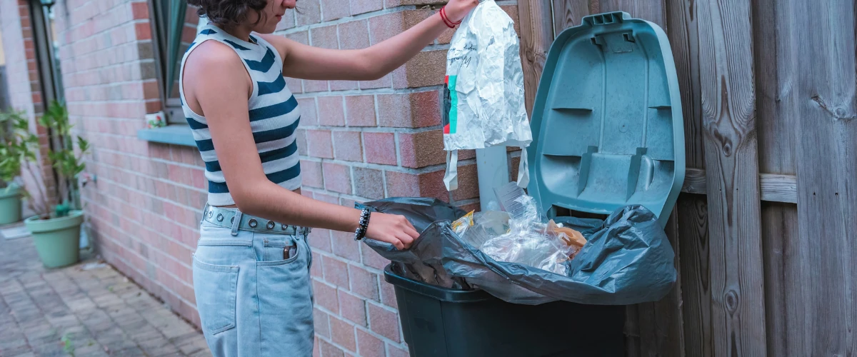 Person placing household trash into an outdoor garbage bin beside a house