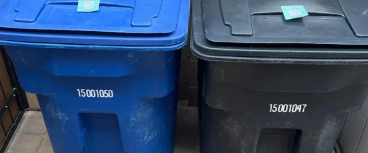 A close-up view of a blue and a black garbage bin side-by-side, each with a QR code sticker on the lid. A close-up view of a blue and a black garbage bin side-by-side, each with a QR code sticker on the lid.