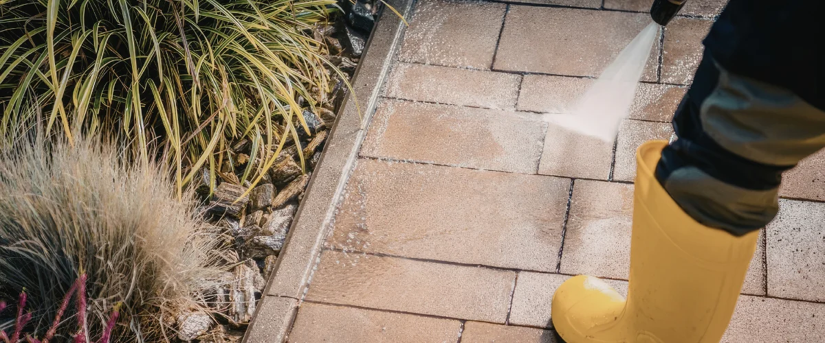 A person in yellow boots pressure washing concrete patio tiles next to a garden bed with green and brown plants. A person in yellow boots pressure washing concrete patio tiles next to a garden bed with green and brown plants.