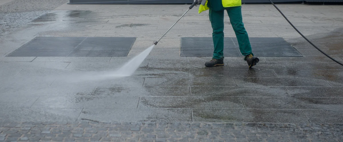 A person in a yellow vest and green trousers pressure washing a wet, grey paved outdoor area. A person in a yellow vest and green trousers pressure washing a wet, grey paved outdoor area.