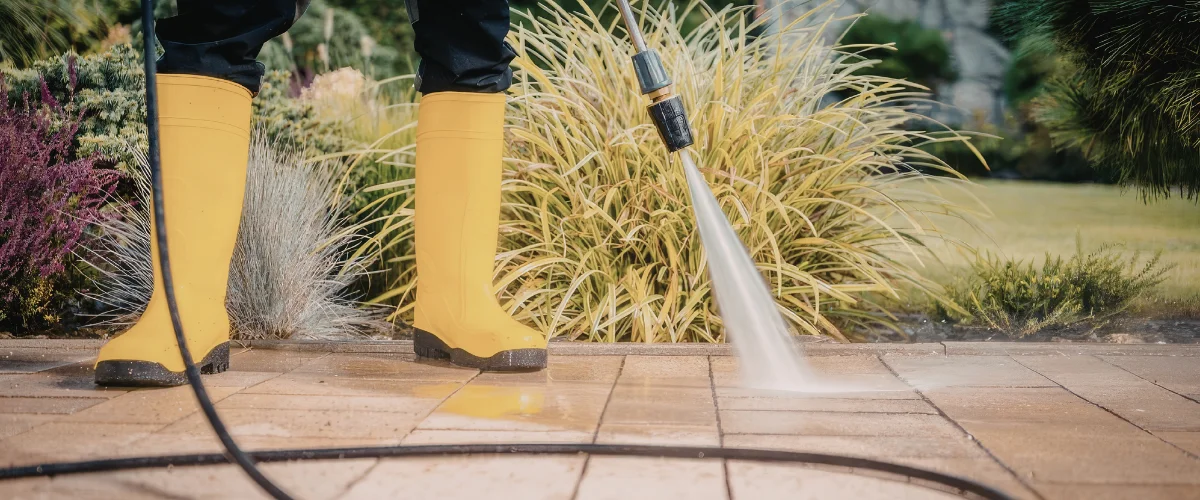 A person in yellow boots power washing a stone patio, with green and purple foliage in the background. A person in yellow boots power washing a stone patio, with green and purple foliage in the background.
