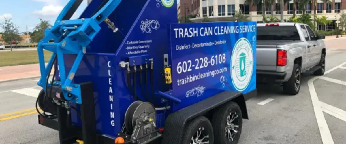 A bright blue trash can cleaning service trailer with a silver pickup truck hitched to it on a paved street. A bright blue trash can cleaning service trailer with a silver pickup truck hitched to it on a paved street.