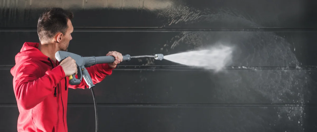A man in a red hoodie pressure washing a black garage door or siding, viewed from the side. A man in a red hoodie pressure washing a black garage door or siding, viewed from the side.