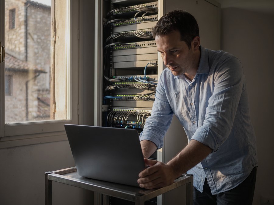 Un professionista dell'informatica al lavoro davanti a un armadio di rete, con il portatile appoggiato su un carrello