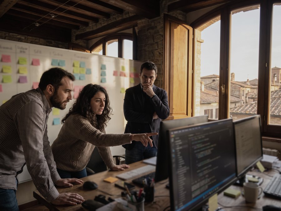 Tre persone di una software house e agenzia davanti a una parete di appunti e a due monitor con del codice