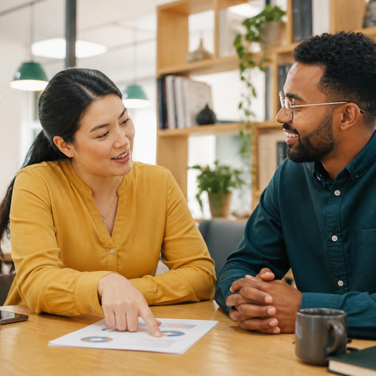 Two people sitting across a table having a relaxed conversation, one pointing to a printed page with simple charts, bright modern office space, photorealistic, approachable and collaborative mood, warm tones Two people sitting across a table having a relaxed conversation, one pointing to a printed page with simple charts, bright modern office space, photorealistic, approachable and collaborative mood, warm tones