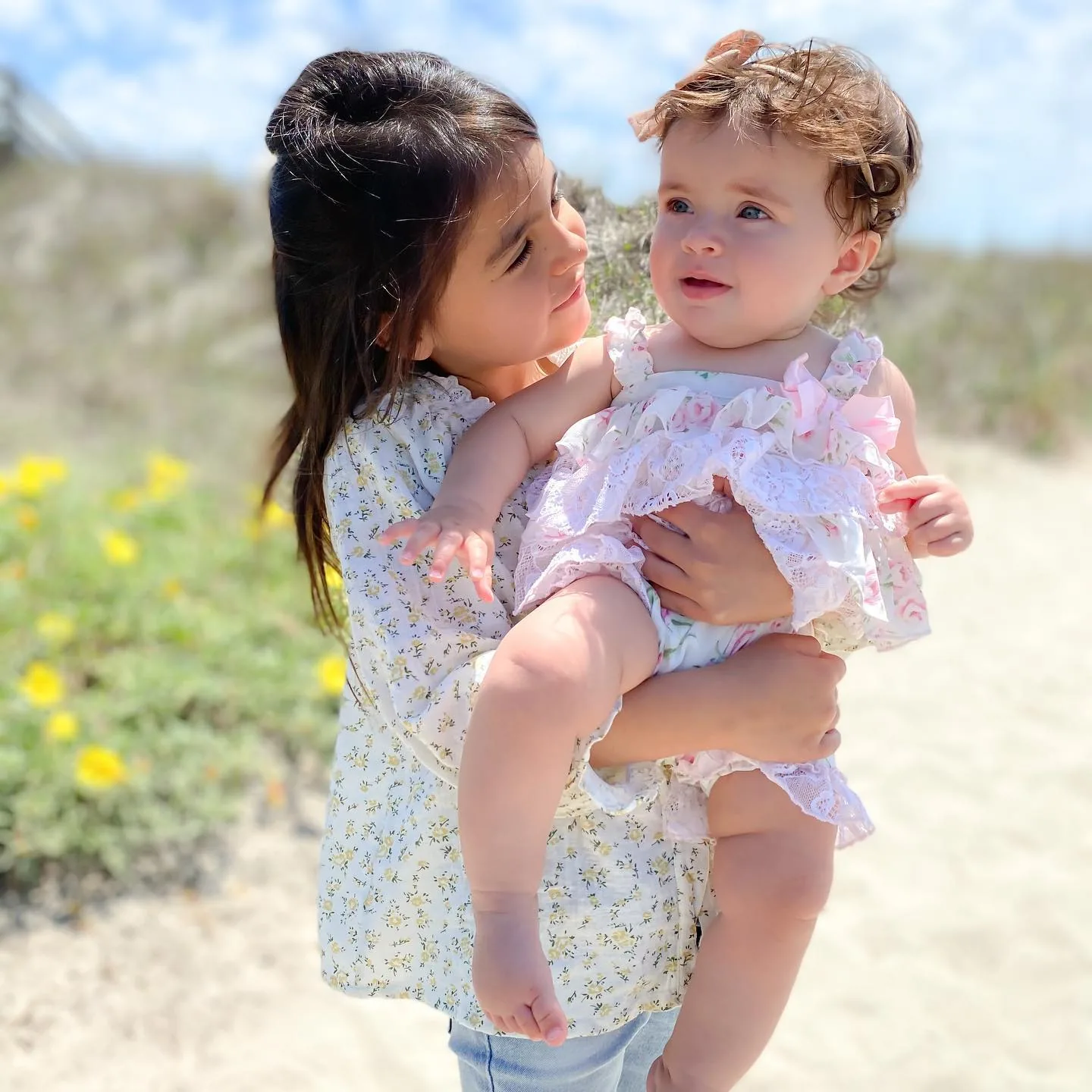 Older daughter holding baby sister in a ruffled dress at the beach