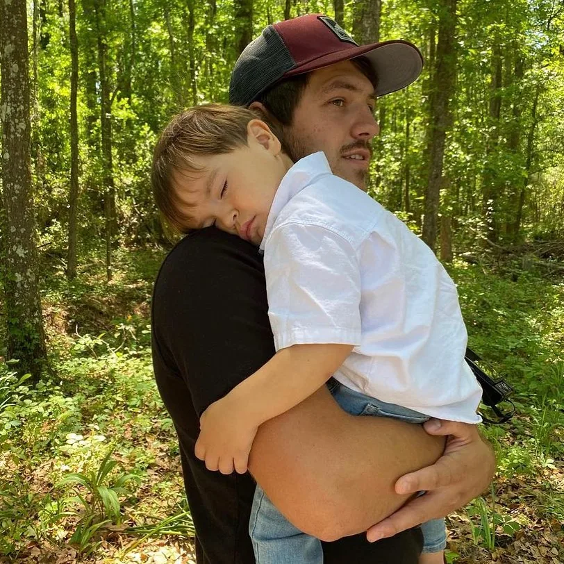 Tanner carrying his sleeping toddler son through the green forest