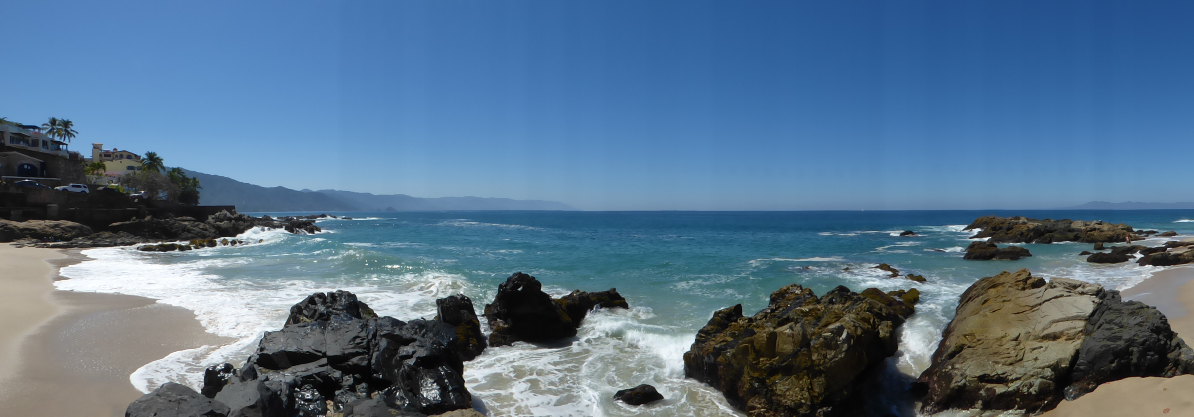 Panoramic view of Conchas Chinas Beach near Puerto Vallarta, Mexico, showing rocky coastline, turquoise Pacific waves, and distant coastal mountains under a clear blue sky.