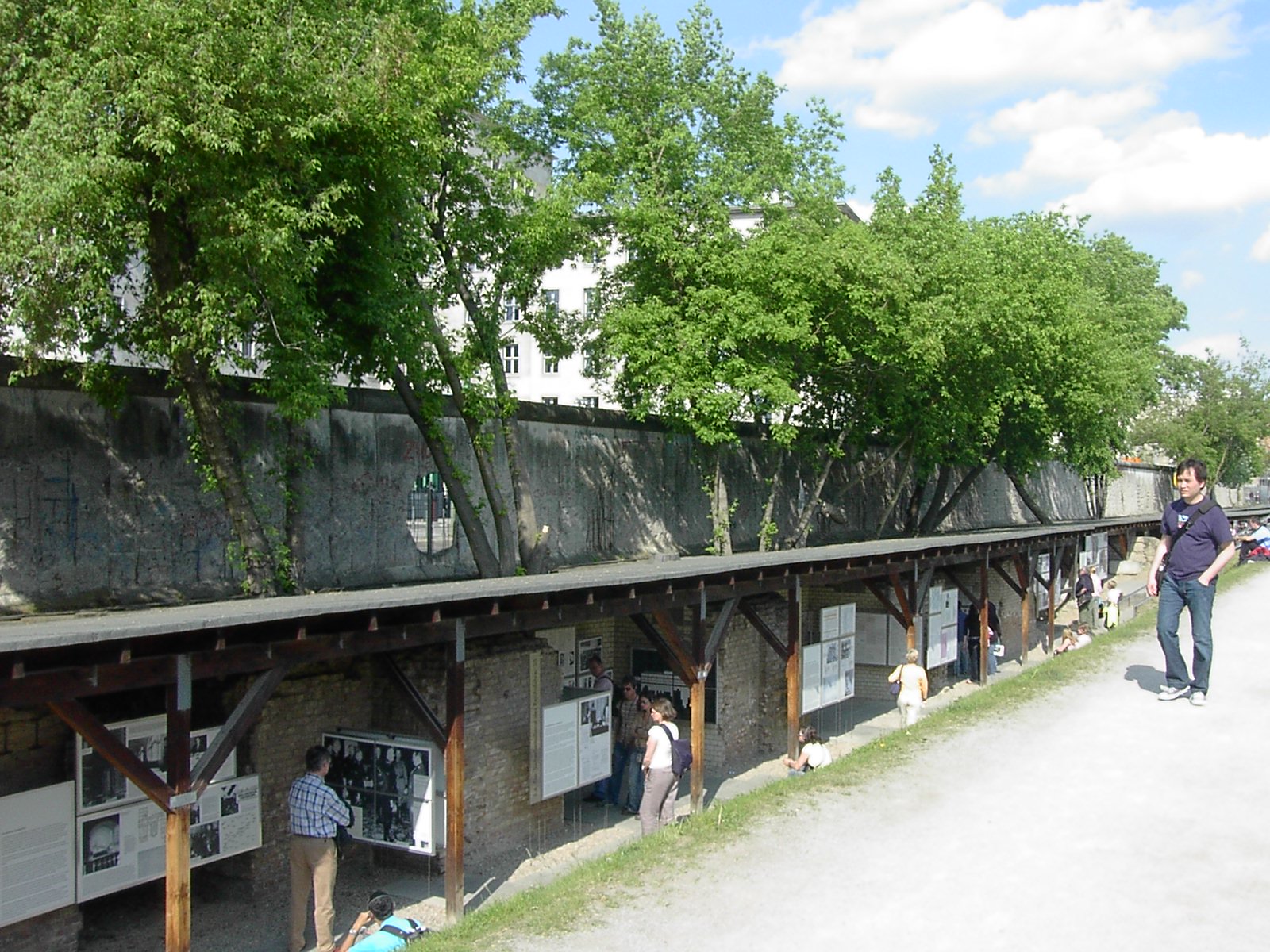 Visitors explore the outdoor exhibits at the Topography of Terror Documentation Center in Berlin, with a preserved section of the Berlin Wall visible above the museum displays.
