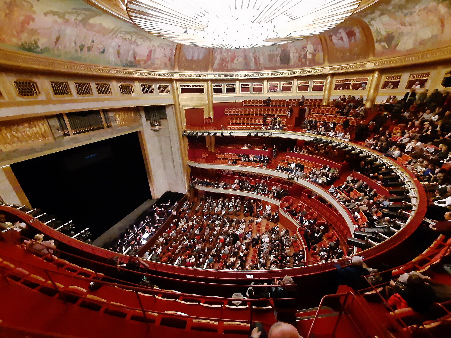 Interior view of the Théâtre des Champs-Élysées in Paris, showing ornate Art Deco balconies, a domed ceiling mural, and a seated audience gathered for a live orchestral performance.
