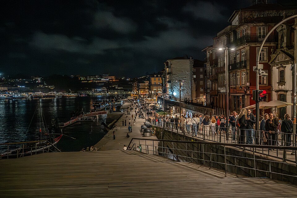 Evening along the Douro River waterfront in Porto, Portugal, with pedestrians, restaurants, historic buildings, and boats.