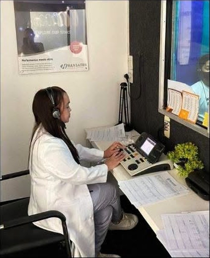 An audiologist working at a diagnostic console inside a hearing test booth