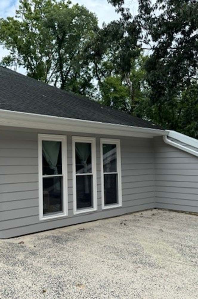 Three tall white double-hung windows set in gray lap siding, with a white gutter and downspout along the roof edge