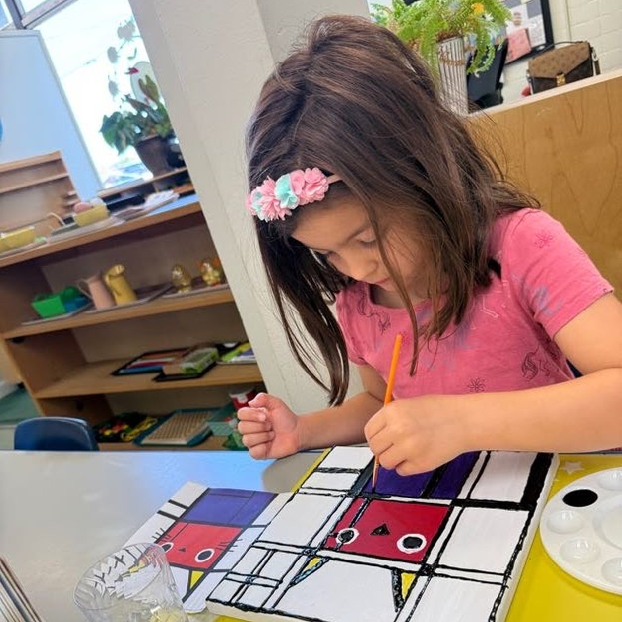 A girl in a pink shirt and flower headband paints a Mondrian-style cat picture on a canvas with a thin brush at a classroom table.
