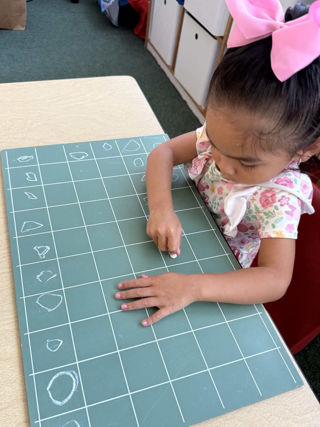 A girl with a pink bow draws shapes in chalk on a green grid board at a classroom table