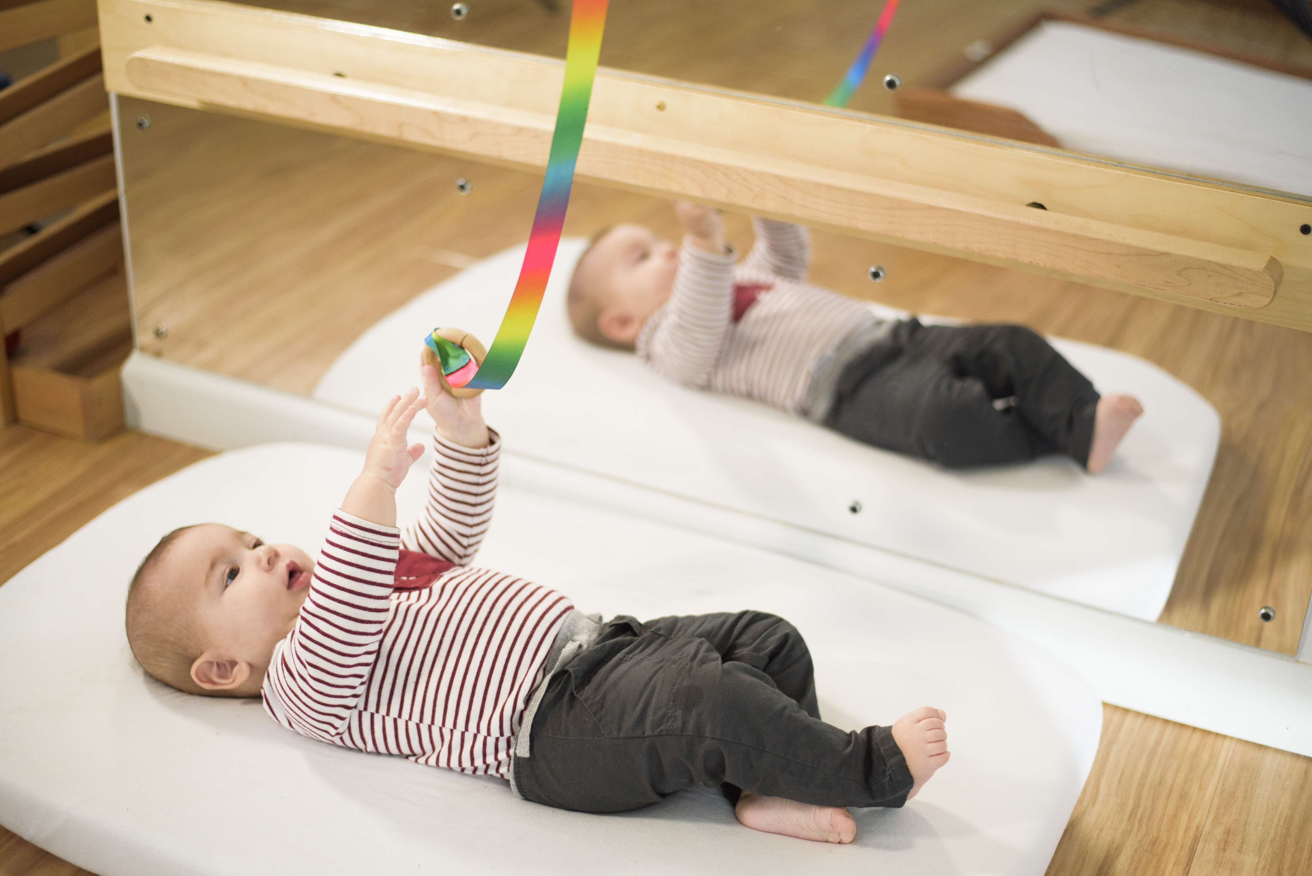 An infant lays on a floor bed next to a wall mirror and pull up bar, reaching for a rainbow ribbon