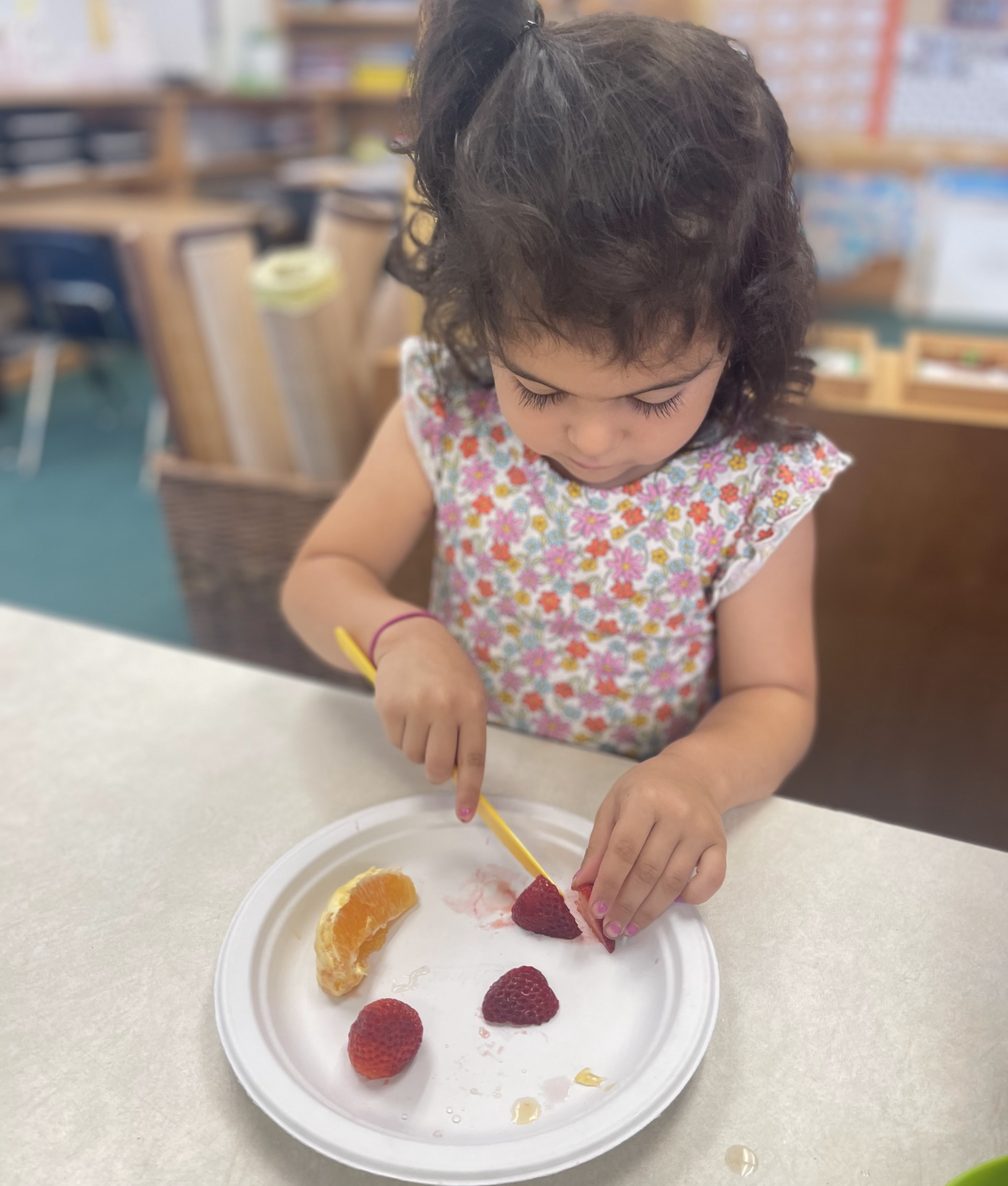A toddler spears fruit from her plate with a wooden pick at snack time