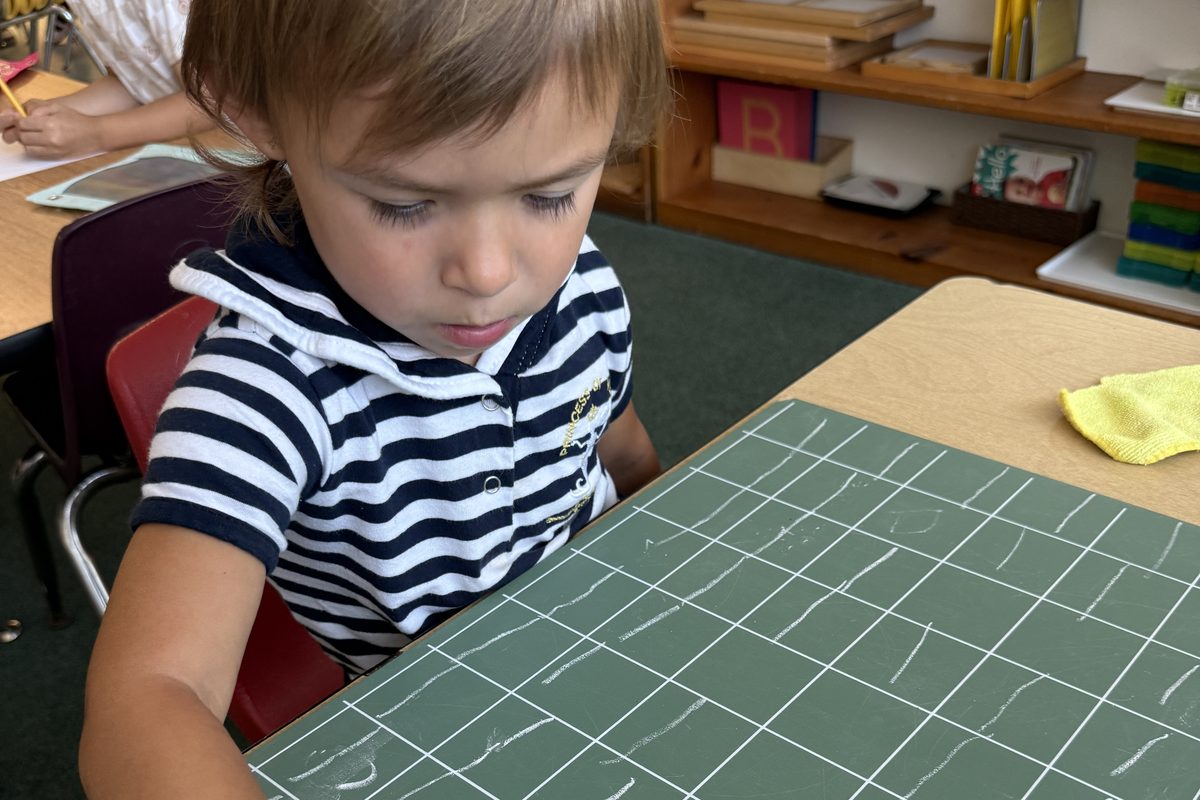 A boy in a striped shirt practises writing on a green chalk grid board
