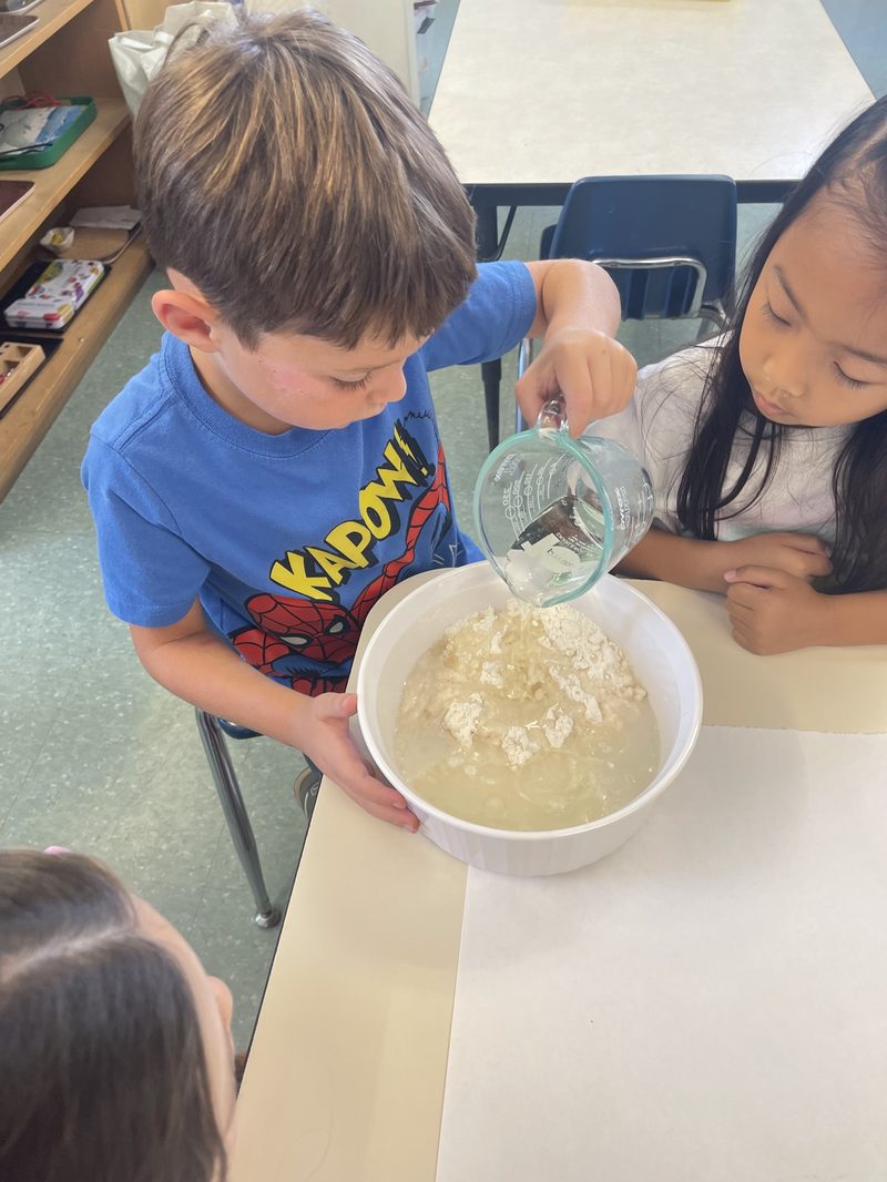 A boy pours water into a mixing bowl while a classmate watches, at a classroom table