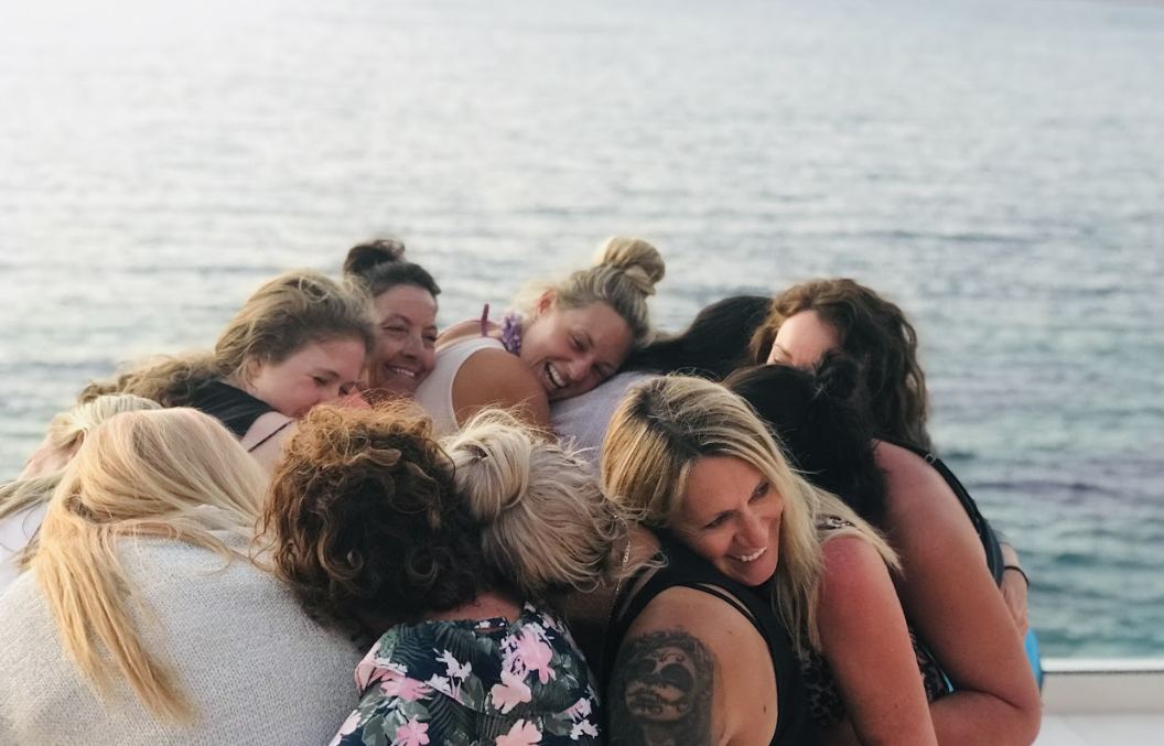 A circle of women laughing in a group hug by the sea