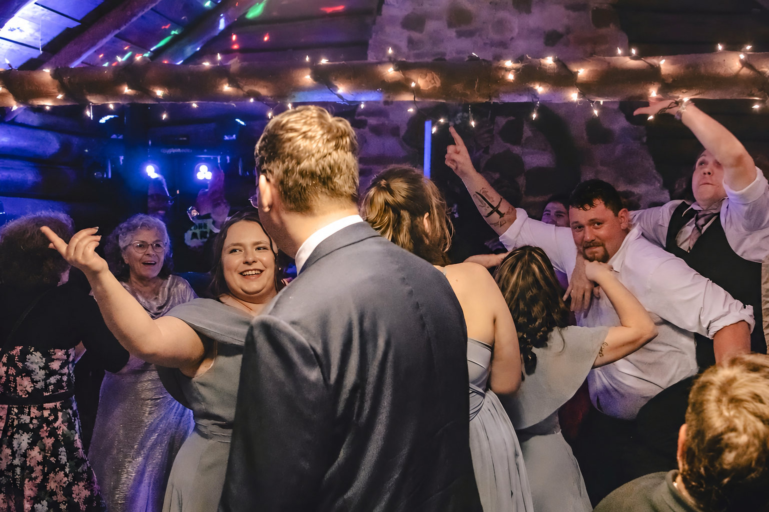 A packed wedding dance floor: the bride, her friends, and her grandmother all dancing together under string lights