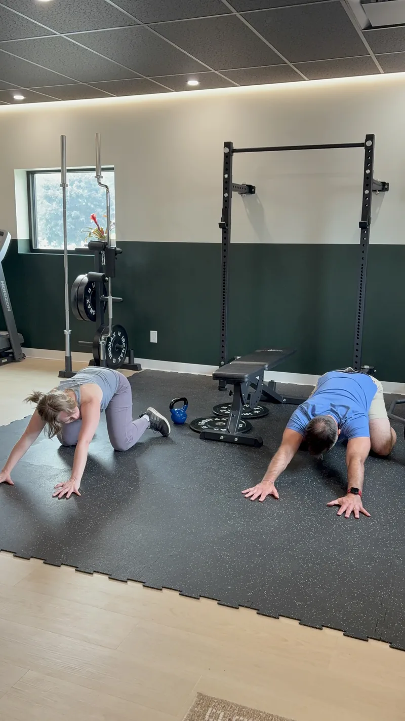 Two patients practicing a floor exercise in the rehab gym at Achieve Movement Health Clinic in Simcoe, with the squat rack and treadmill behind them