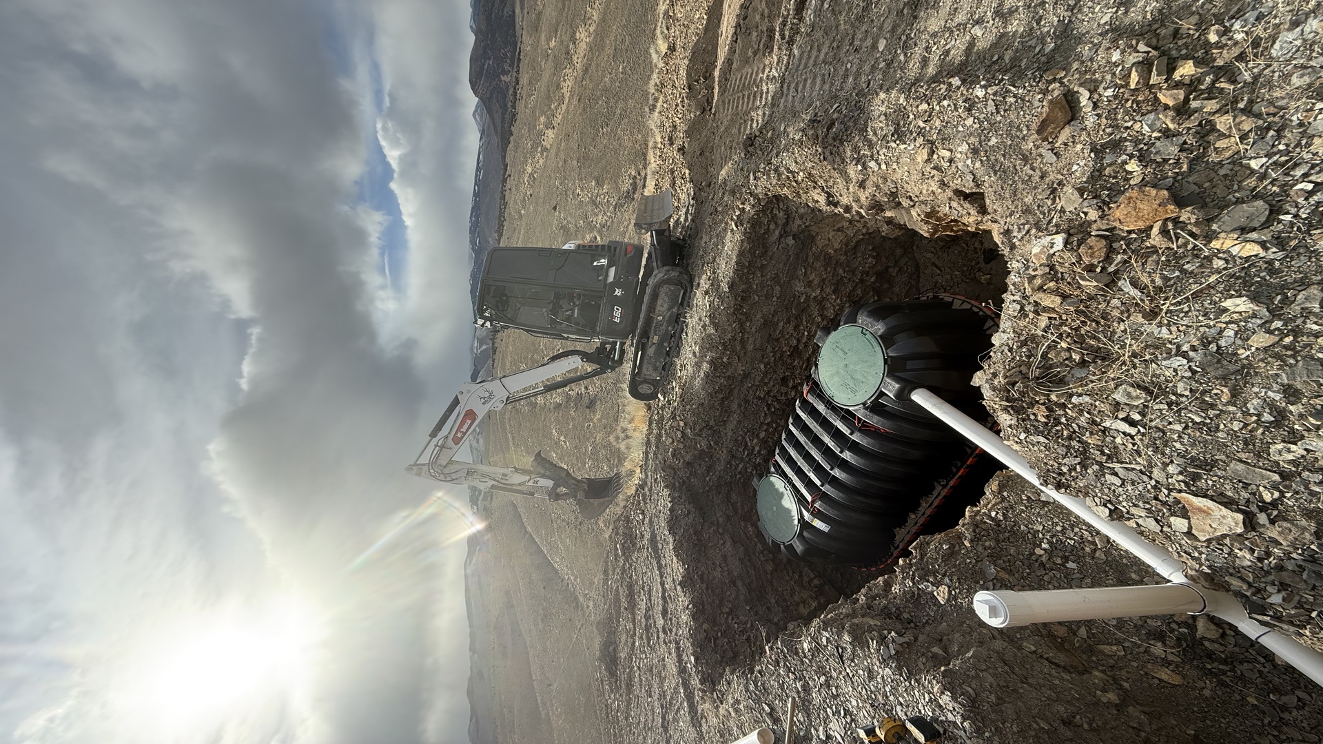 Excavated septic leachfield trenches on a rural Fremont County, Wyoming property, showing gravel bed placement and leveled trench walls ready for pipe installation. Fremont County high desert terrain visible in the background.