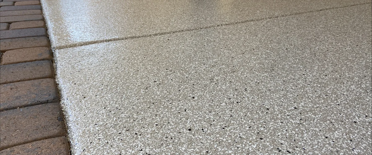 Close-up of tan speckled epoxy garage flooring next to a border of brown brick pavers.