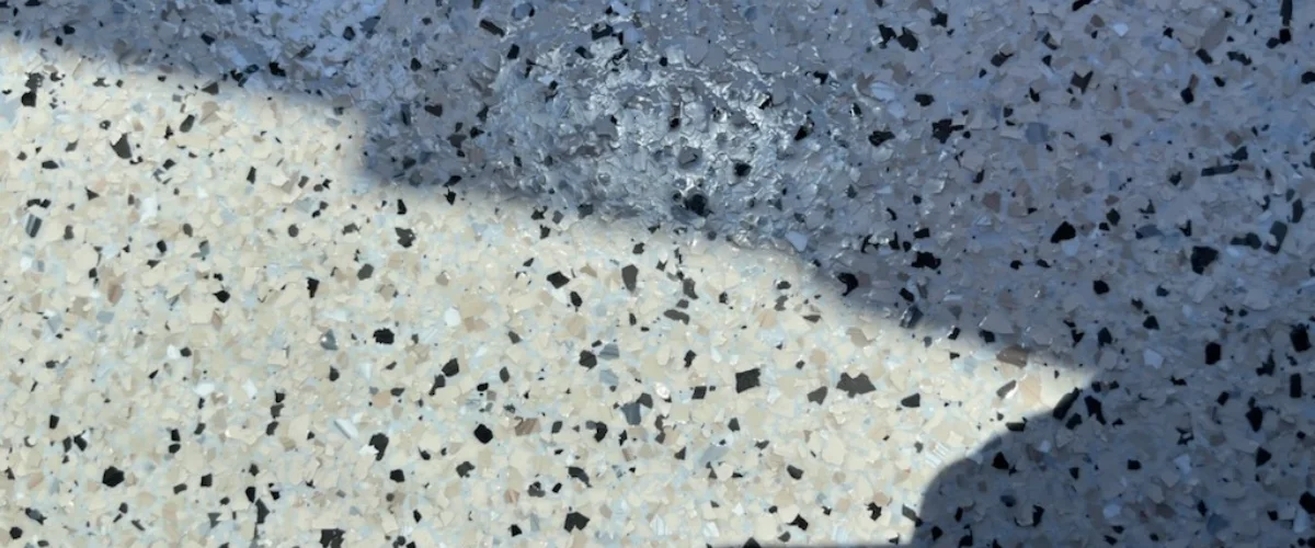 A close-up view of light-colored granite-like epoxy flooring with dark speckles and a reflective sheen.