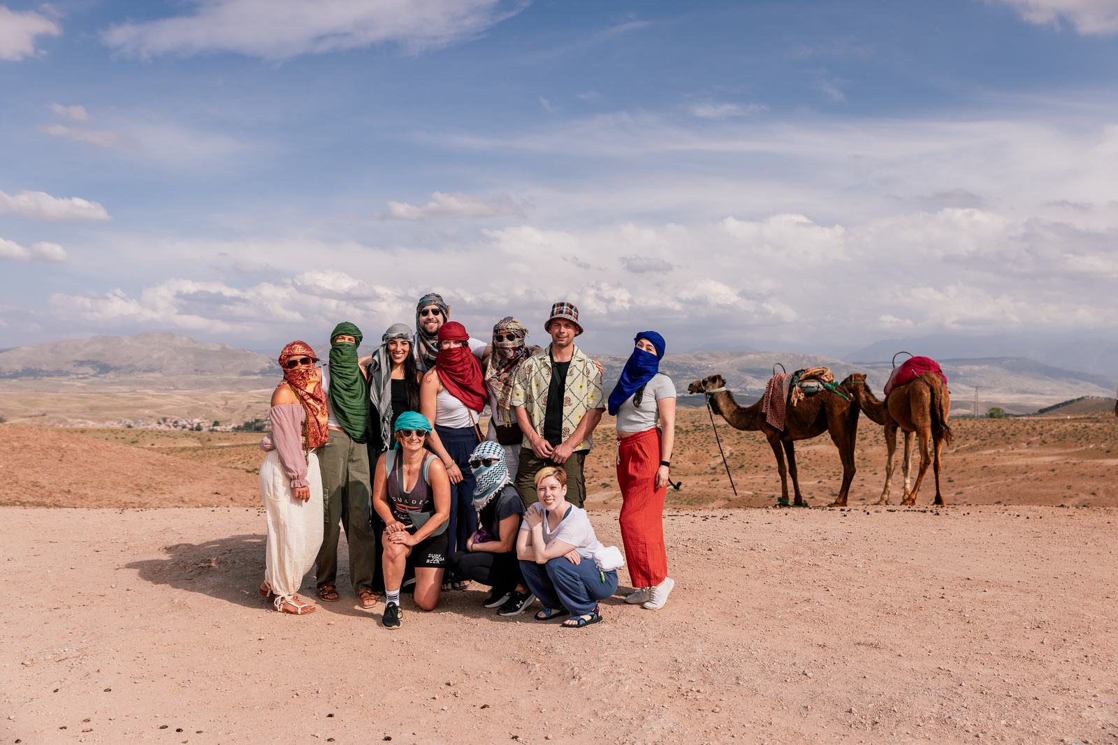 A small group in headscarves standing in the desert beside camels