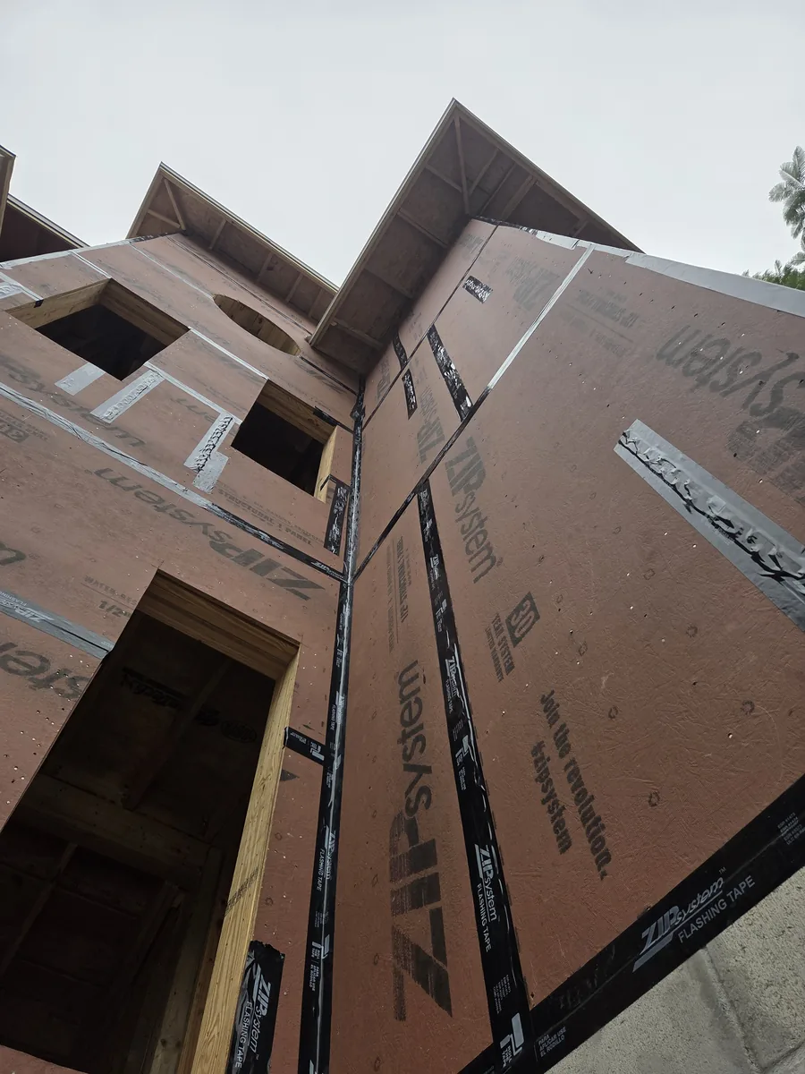 A house under construction, its walls wrapped in sheathing, seen from below against the sky
