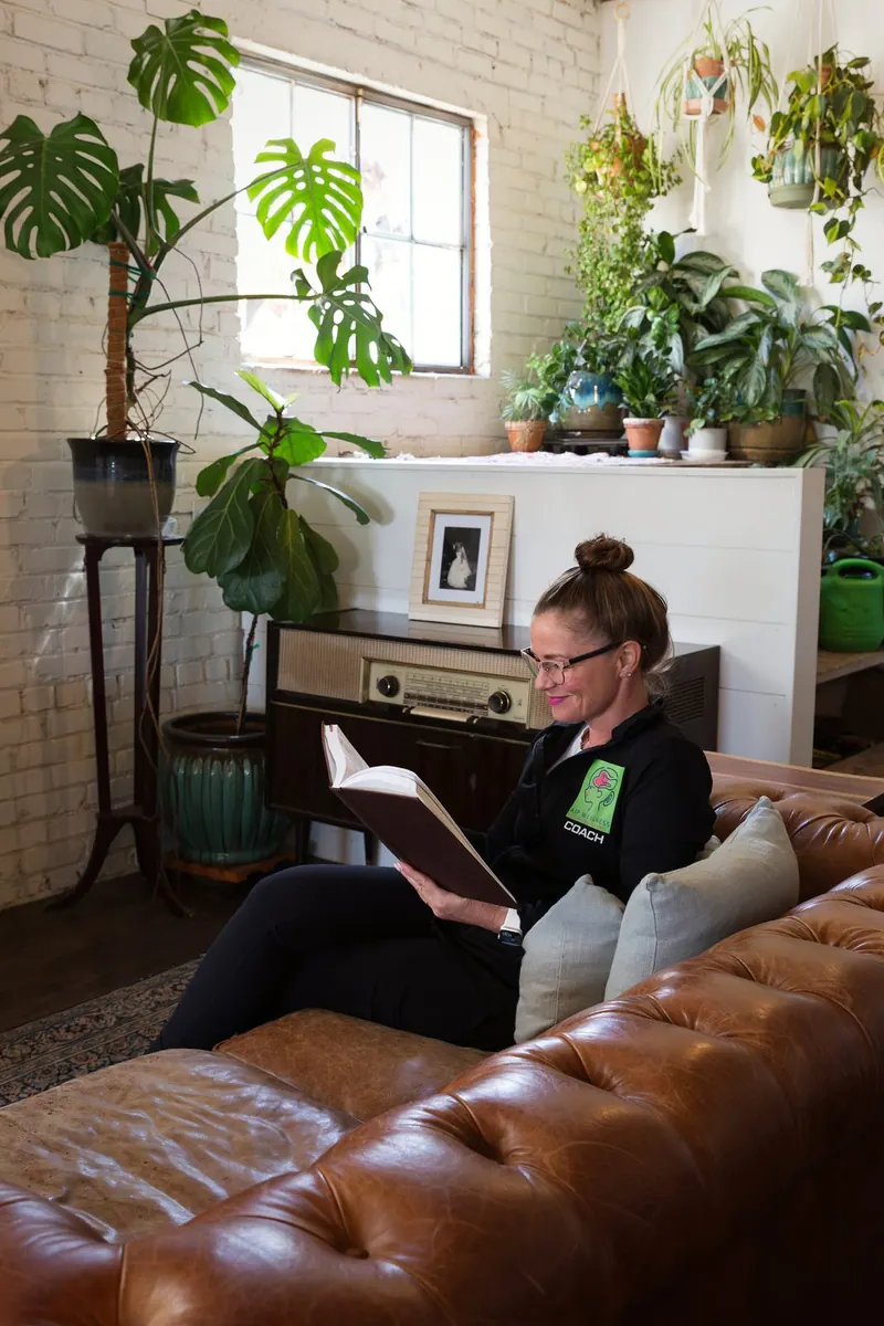 Anya Gossage reading a book on a leather sofa in a plant-filled living room